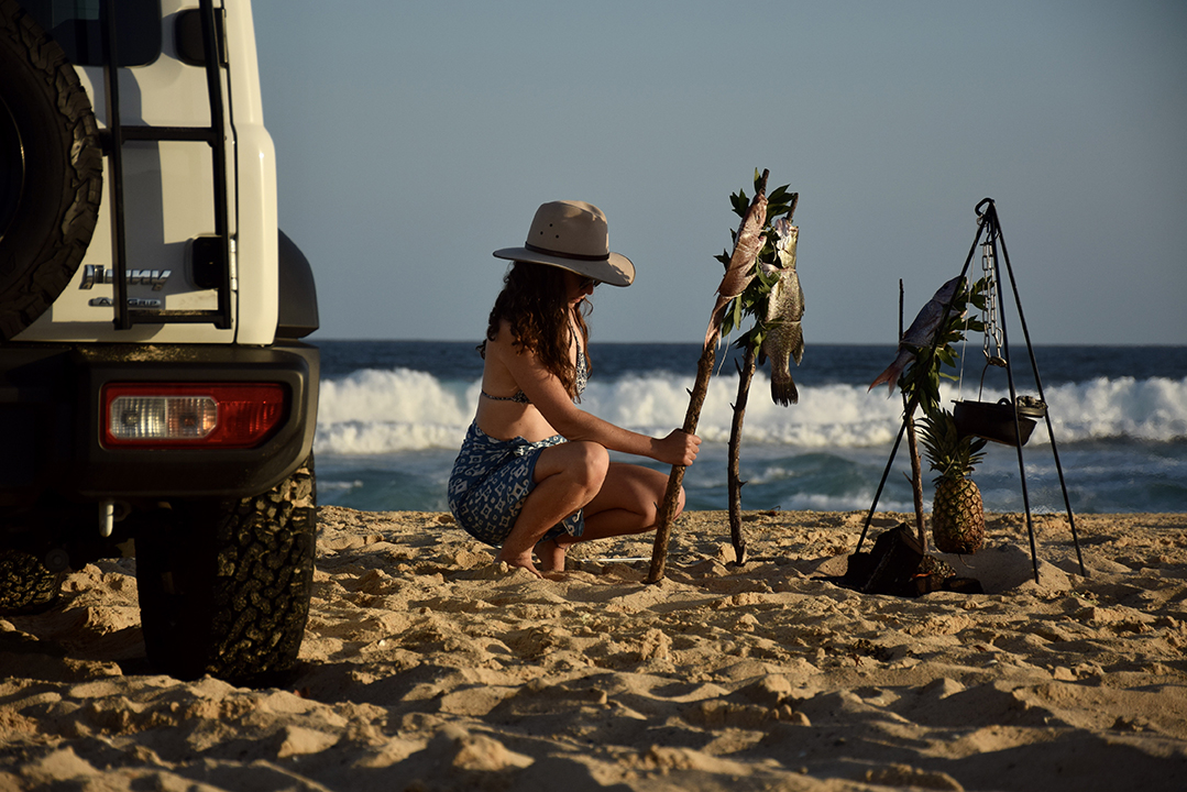 Woman cooks fish on beach next to SUV.