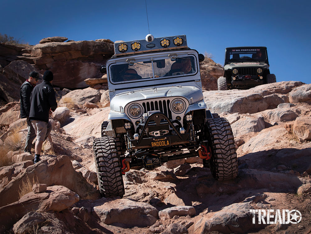 Silver Jeep drives down rocky trail with two spotters off to the left side.