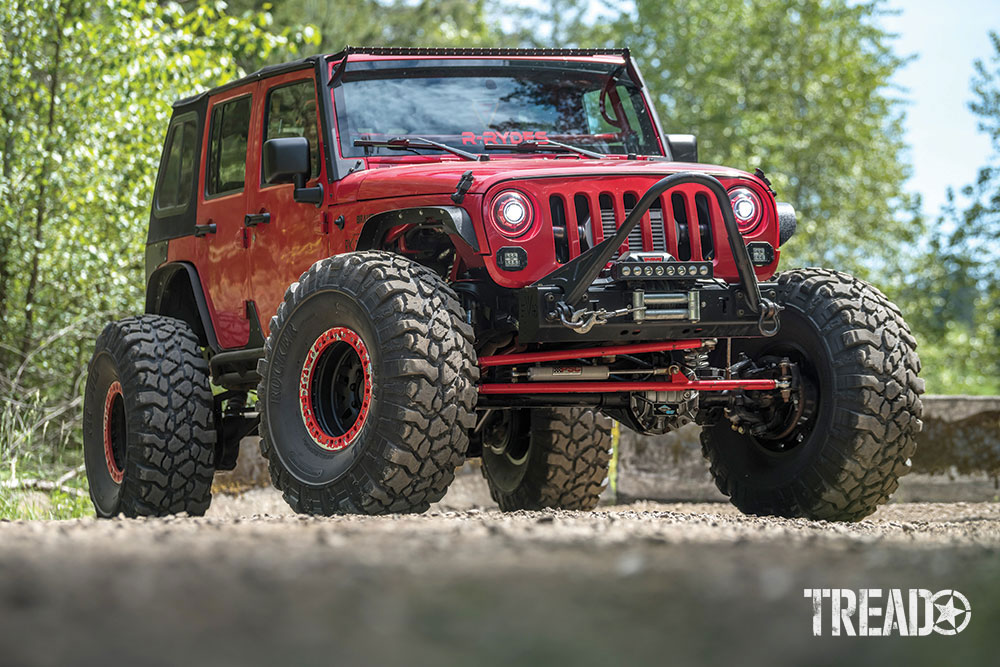 A red Jeep JKU is parked on a dirt road with green trees behind it.