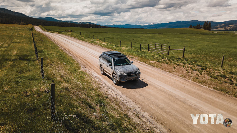 A black Lexus LX570 drives down a dirt road. A roof rack and recover boards are seen mounted to the roof.