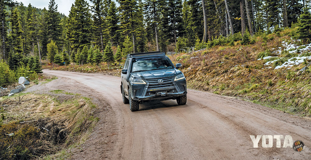 The black Lexus LX 570 drives down a dirt road surrounded by tall trees.