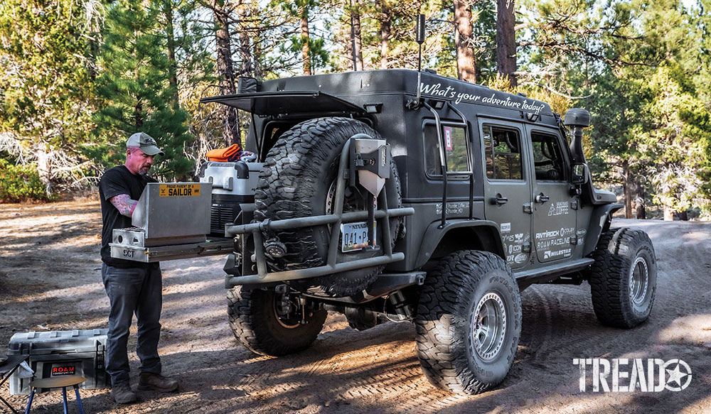 Man cooks on his stove that slides out from rear of Jeep while parked in mountains.