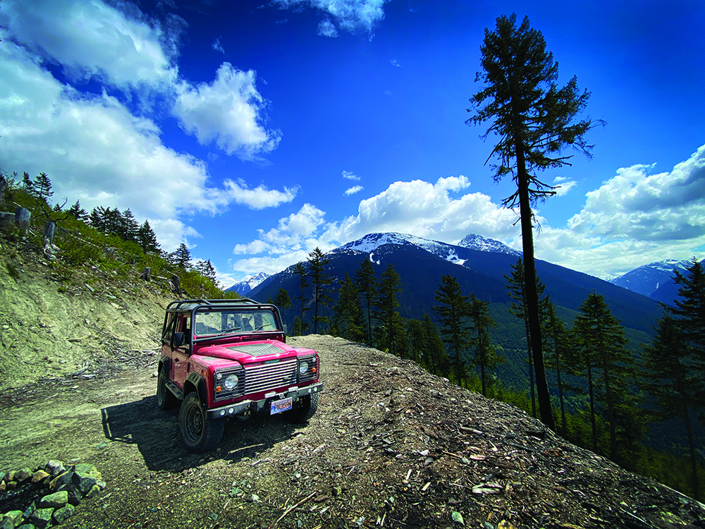 A wide-angle shot of the Defender on a rocky path with blue sky and mountains behind.
