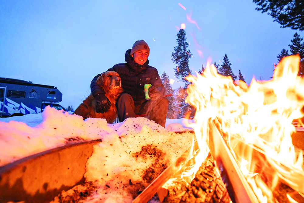 Andrew Muse and his dog, Kicker, sit by the fire at a snowy campsite.