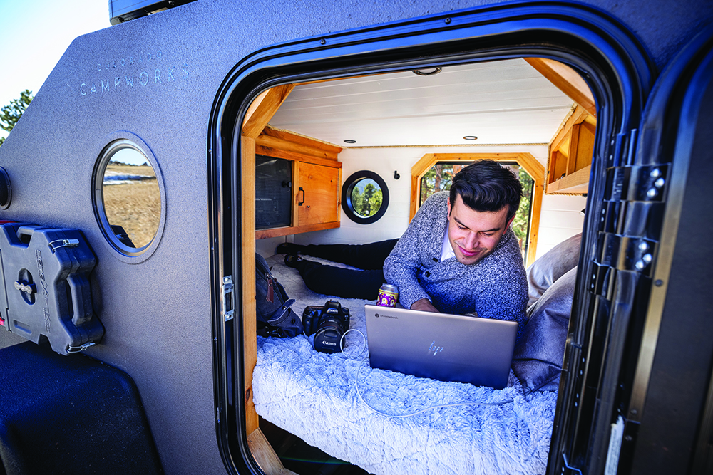 A man looks at a laptop while laying on the mattress inside the camper.