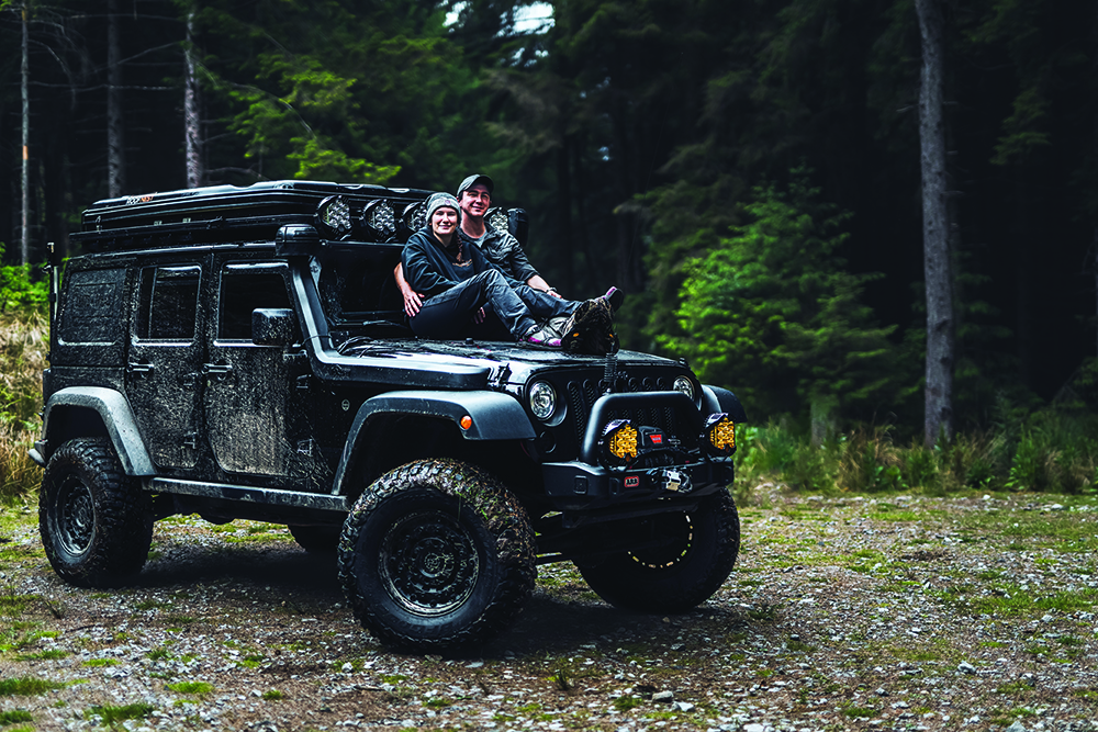 Thomas and Hannah Tucker sit on the hood of a jeep parked in the forest. 