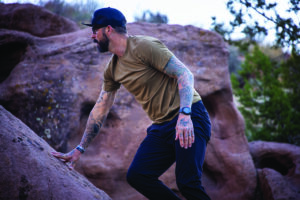 A tattooed man in a green performance tee shirt scales a rock.