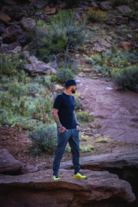 A man in dark clothing and neon shoes poses on rocky background.