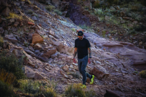 A man walks along rocky terrain in dark clothing and neon shoes.