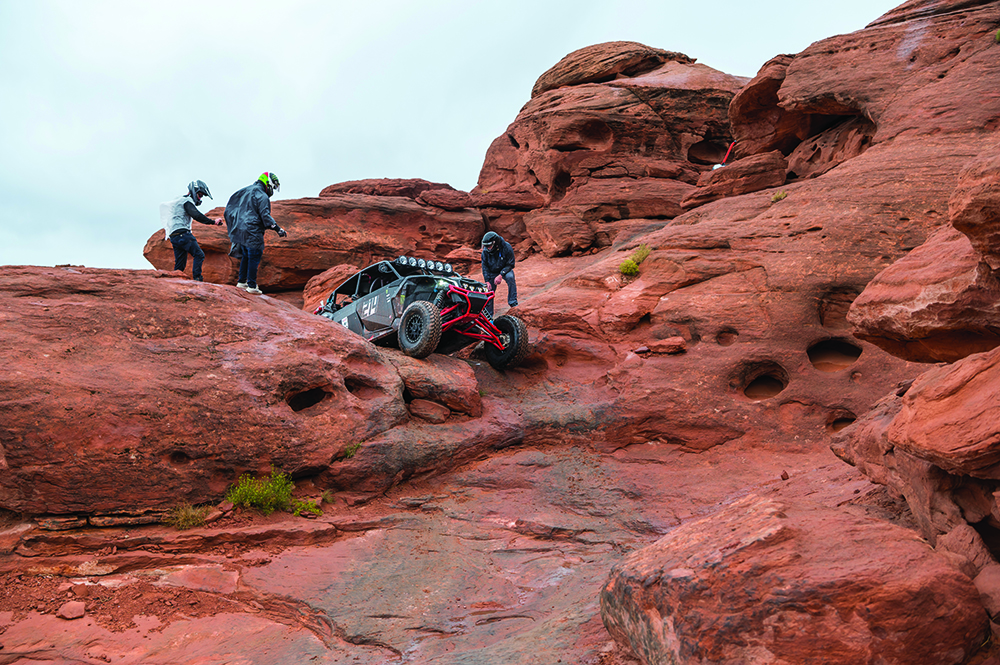 Several competitors in helmets coach a UTV through a red rock obstacle.