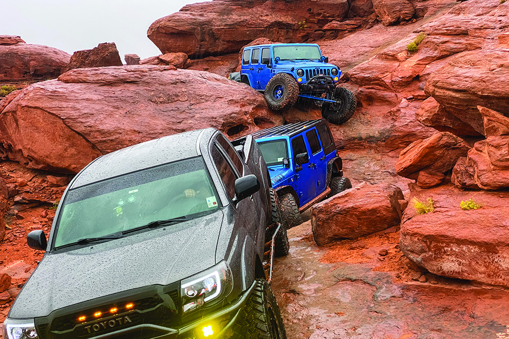 Two blue Jeeps follow a black Tacoma down a steep slope.