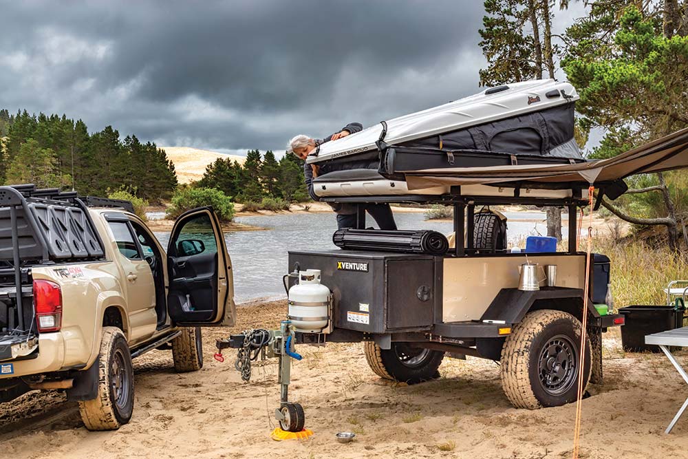 The tan Tacoma truck and trailer park next to a lake on sand.