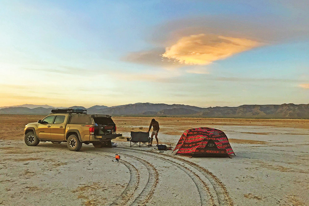 A yellow truck parks on the desert sand with a red tent behind.