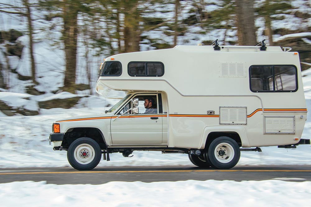The white camper with orange stripe drives down pavement through a snowy wood.