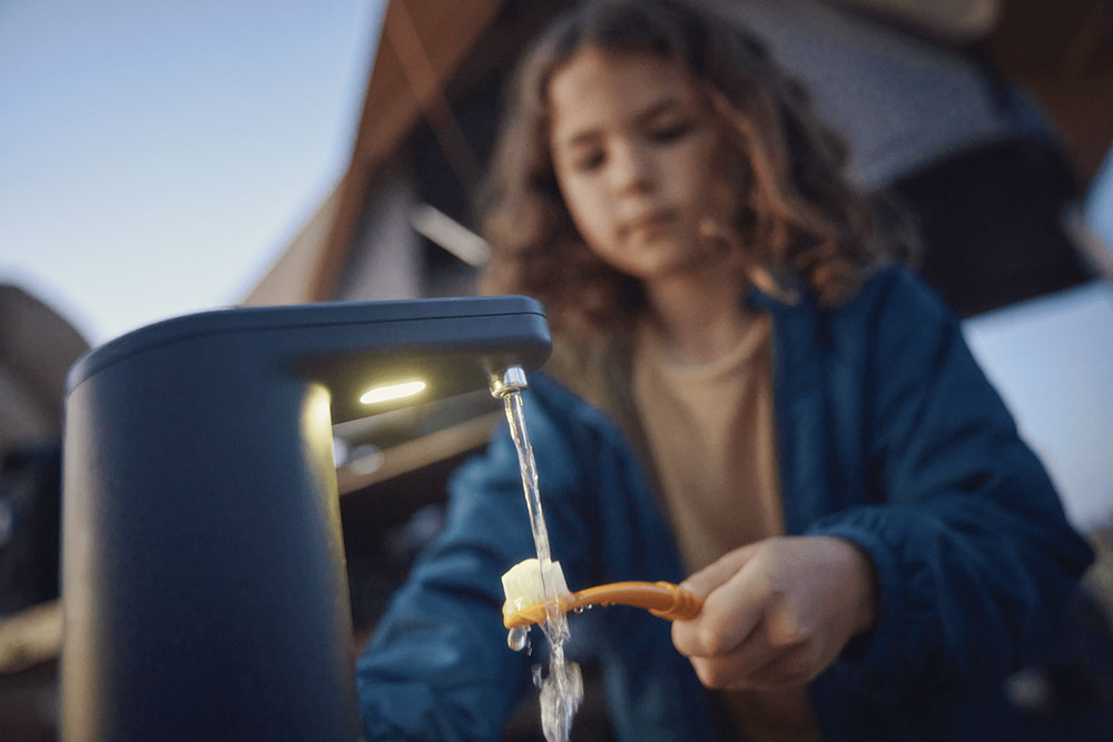 A girl holds her toothbrush under the water from the faucet.