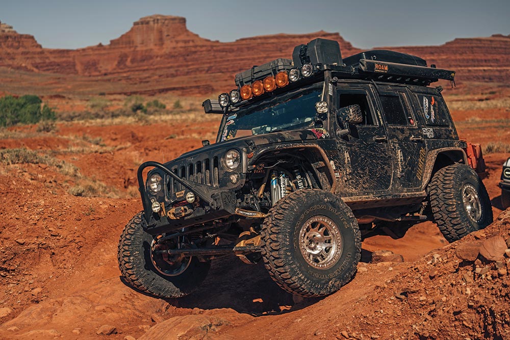 A black jeep drives over red sand and rocks.