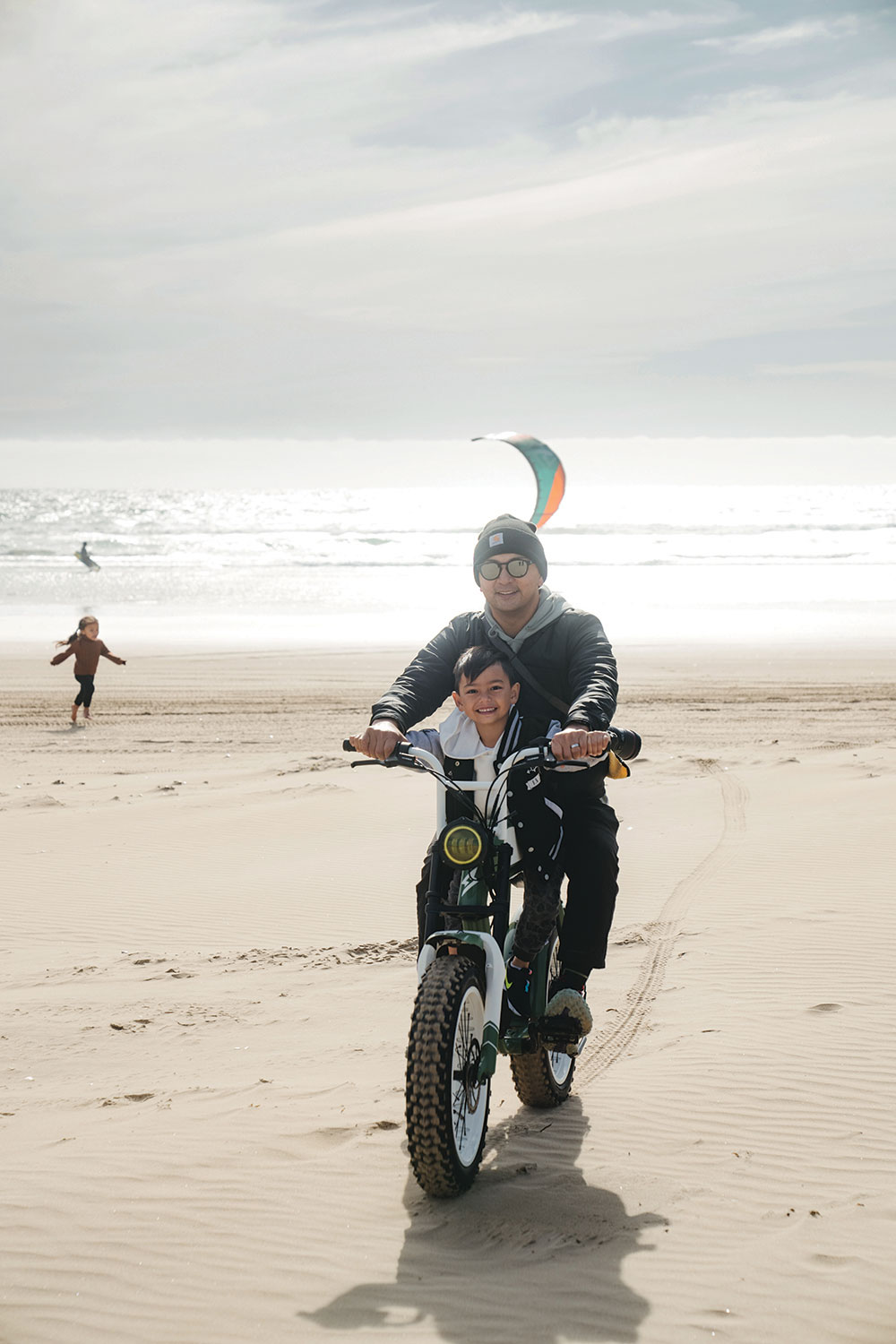 A man and a small child ride an electric bike on the beach.