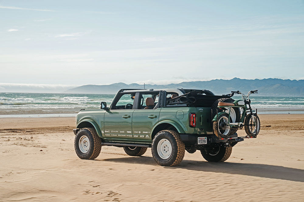 The green Ford Bronco, with bike on the back, parks on the beach with the top down.