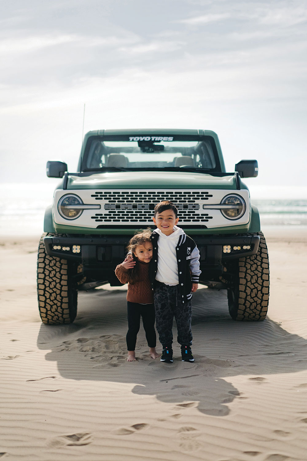 Two small children stand arm in arm in front of the Bronco.