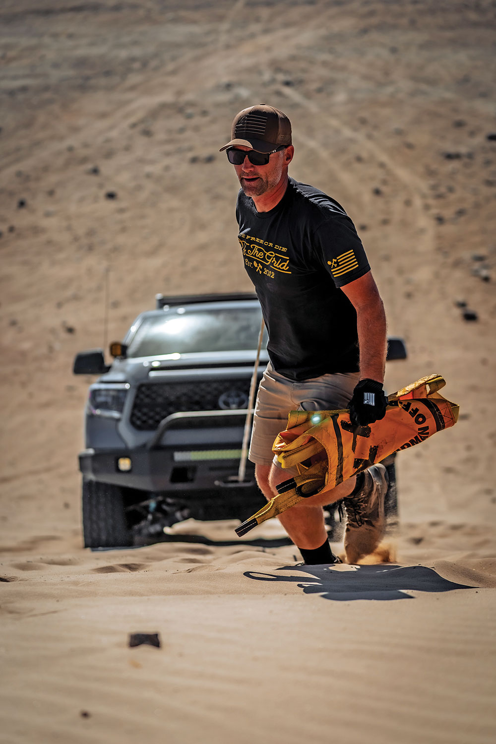 Samuel, in a black shirt and hat, carries a Deadman Offroad recovery anchor uphill from the truck.
