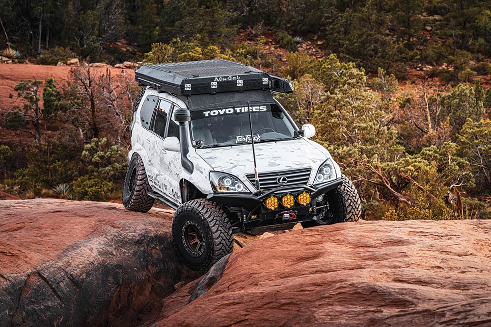 The white GX470 uses its custom suspension to drive over a gap between two red rocks.