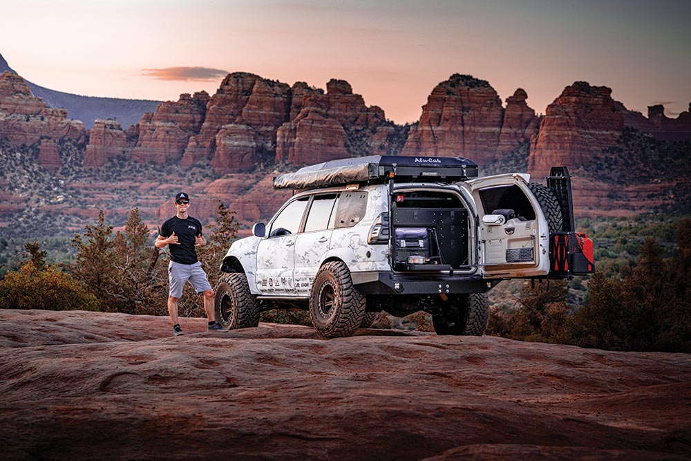 The truck's owner poses next to it with thumbs up in front of stratified rock mountains.