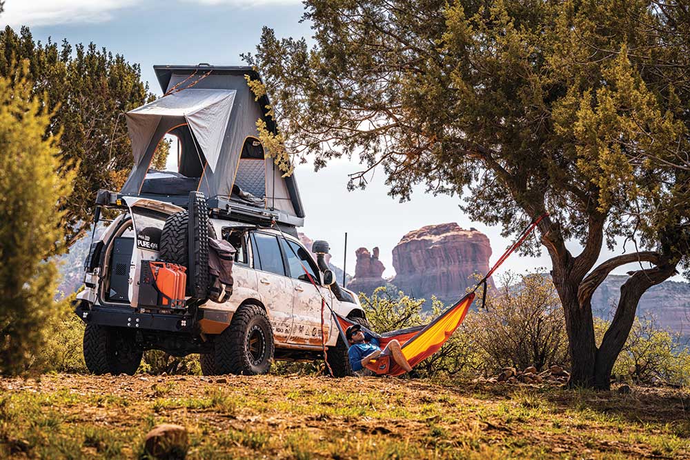 Someone naps in a yellow hammock strung between the GX470 and a tree. The rear door of the truck is open and the roof top tent is deployed.