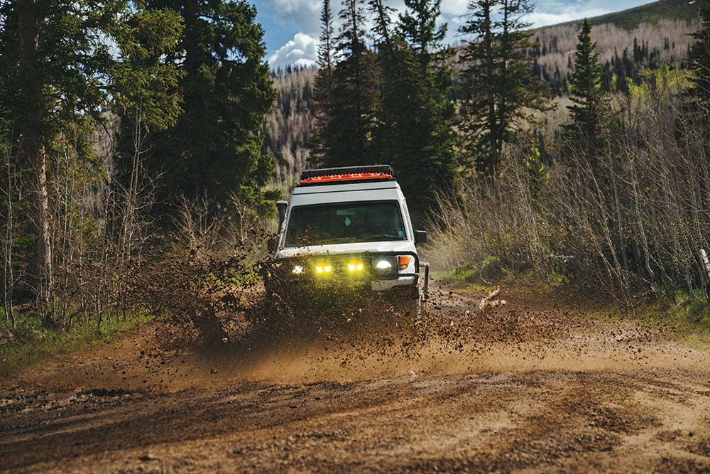 The white Troopy kicks up mud while traveling through a forest of evergreen trees.