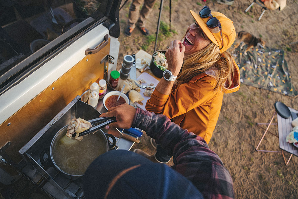 A man and woman cooking on the tailgate of the SUV while traveling. 