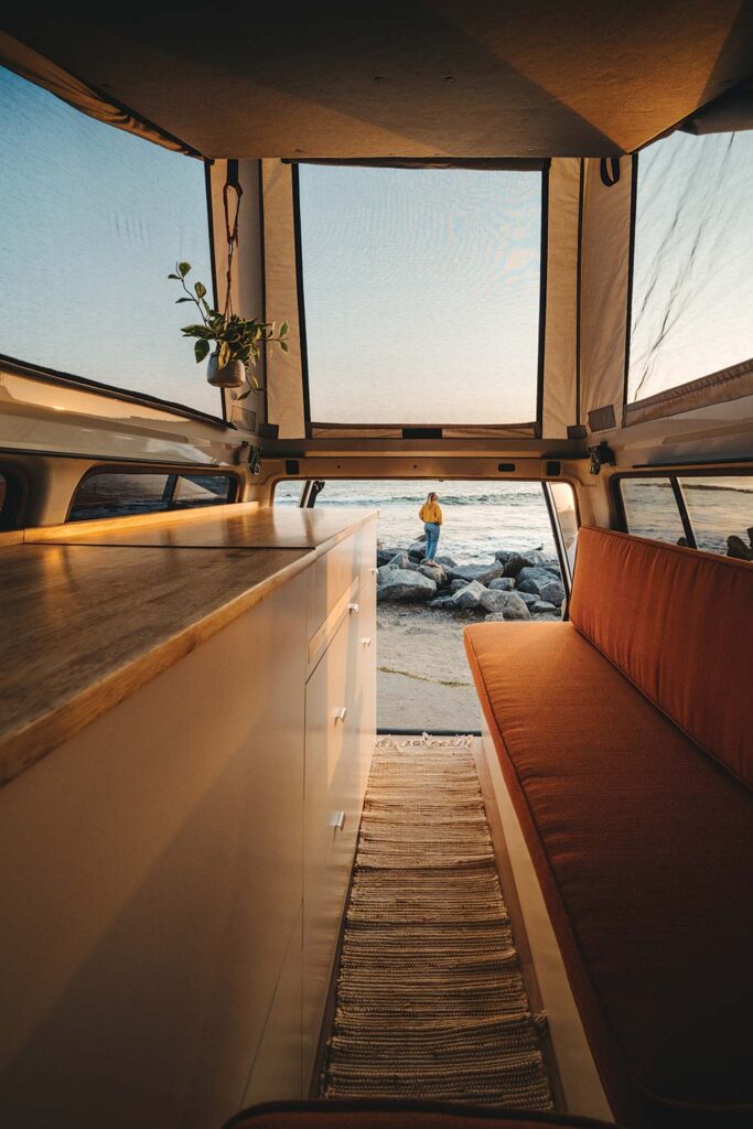 The view of a man standing by the ocean from the inside of the Troopy, showing the countertop and bench.