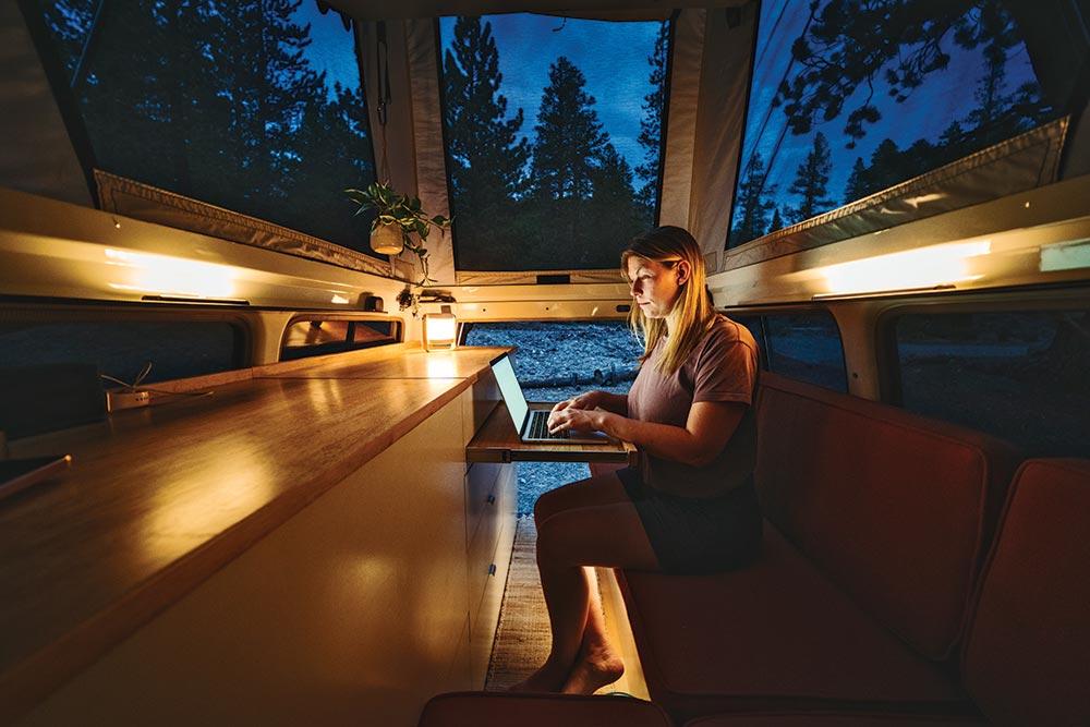 A woman sits on the bench in the back of the Troopy working on her laptop while traveling.