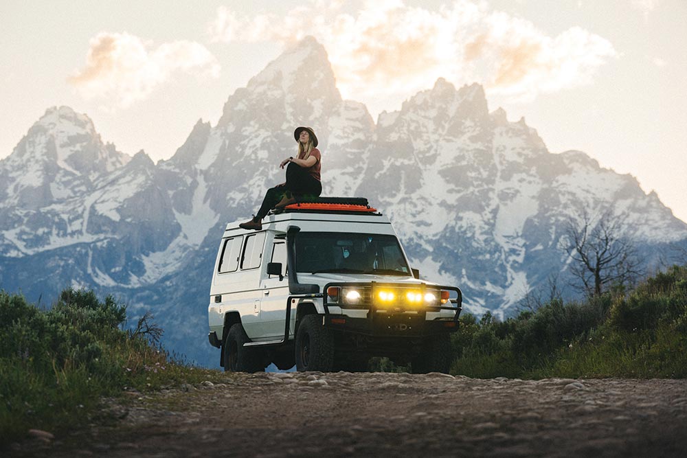 A woman sits on top of the white SUV while traveling in front of a huge mountain.