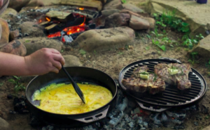 A person stirs eggs in one cast iron pan while steaks rest on another at a camp.