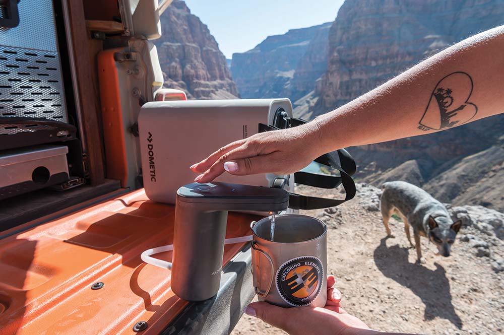 A hand reaches into the frame to tap the top of a faucet that dispenses water into a metal cup.