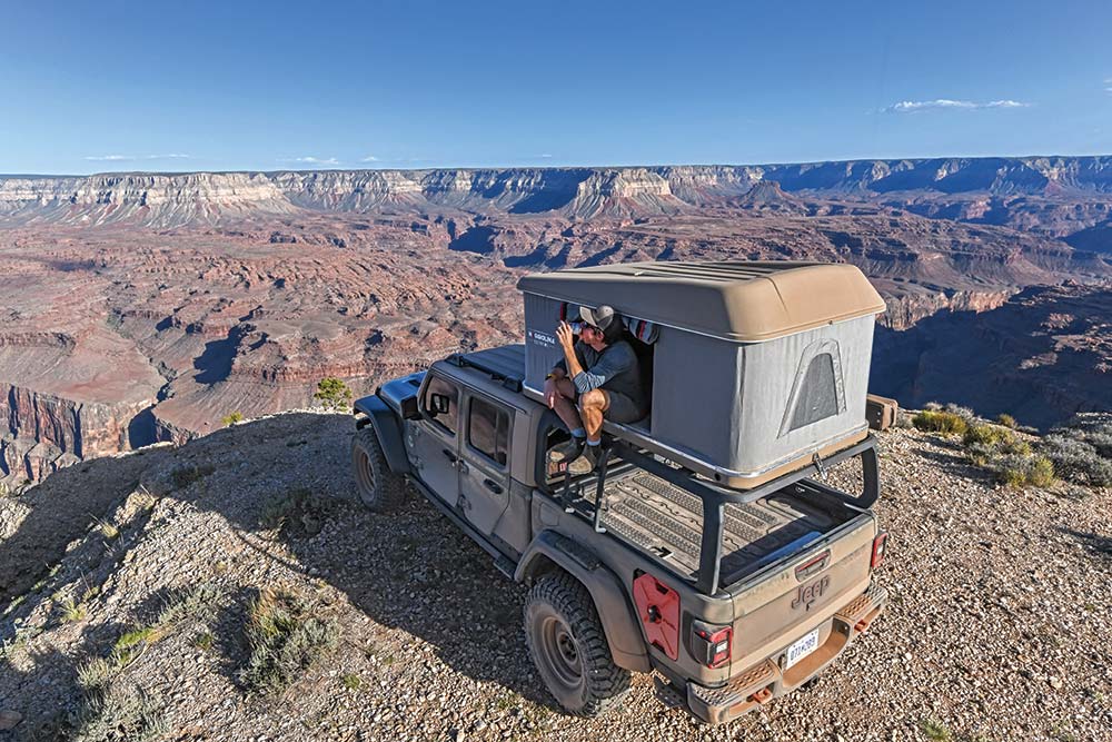 Man sits on edge of rooftop tent on top of Jeep Gladiator and enjoys a drink while looking out at the Grand Canyon.