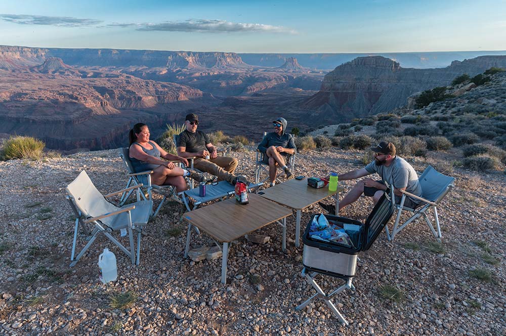 A group of four overlanders sit in camp chairs on a canyon cliff, enjoying the day.