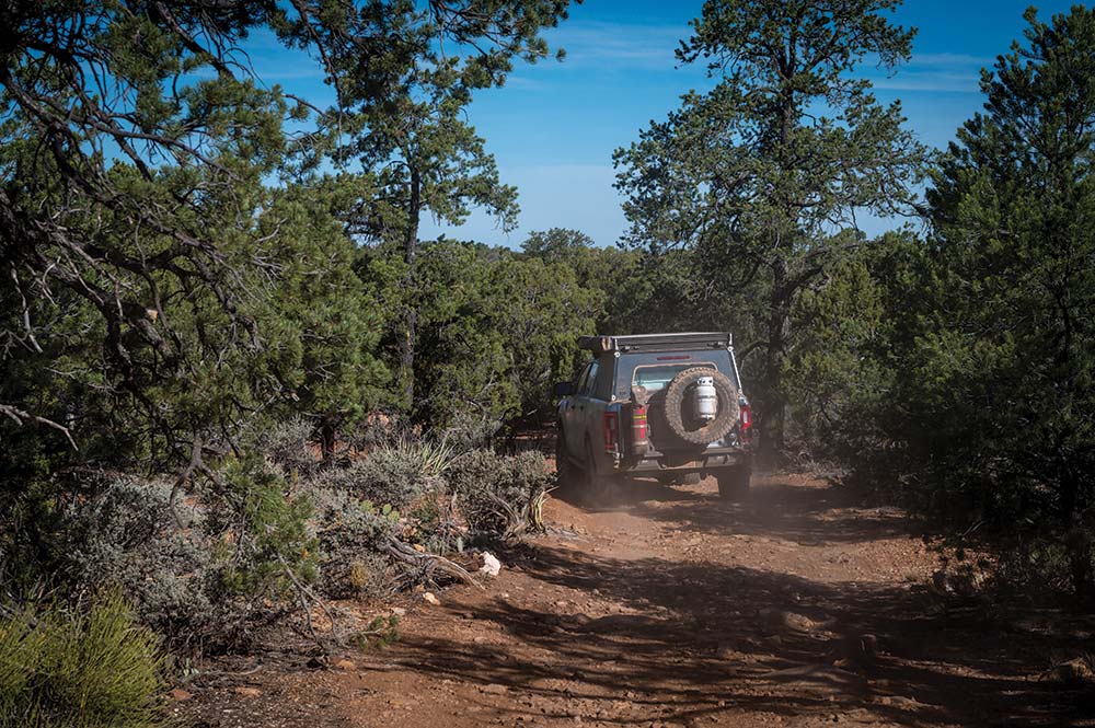 An overlanding vehicle with a large spare tire mounted on the back drives into the woods.