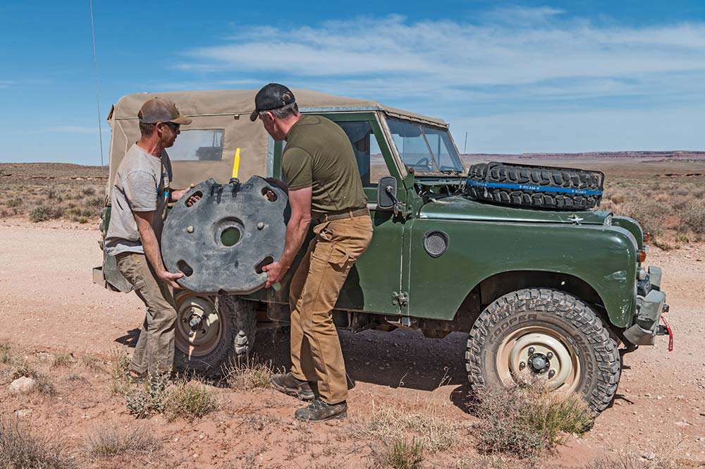 Two men fuel up a vintage green Land Rover Defender with a spare fuel container.