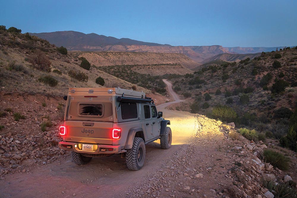 A Jeep Gladiator with bed cover and racks covered in dust uses its headlights to illuminate a dirt trail at dusk.