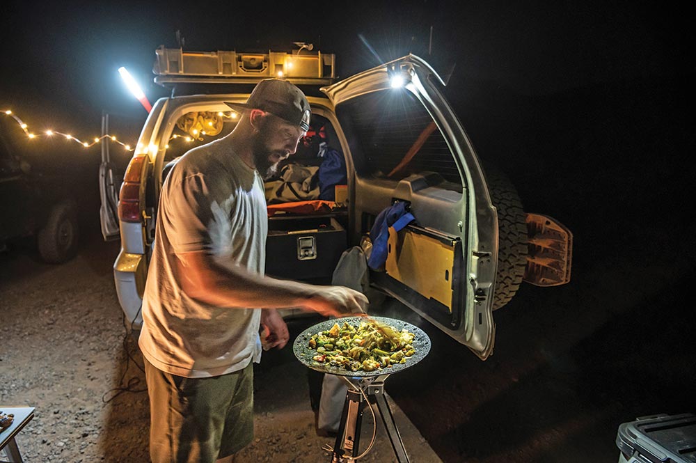 A man in a backwards hat cooks a stir fry in a stand up wok, in front of the open back of his overlanding vehicle.