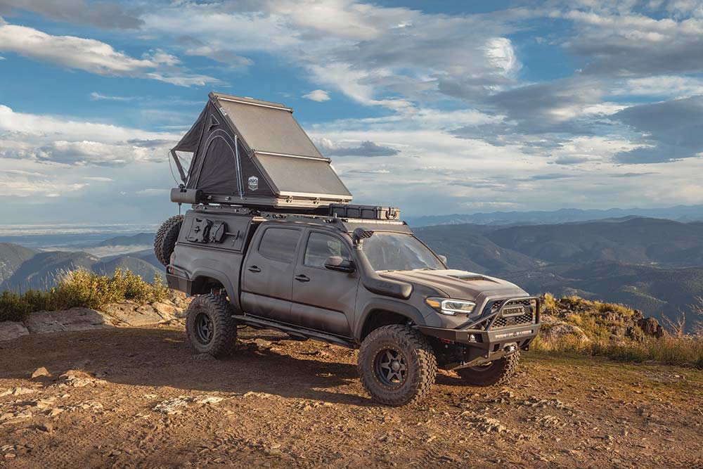 Parked on a cliff overlooking a valley with roof top tent deployed, the HerschX Tacoma is covered in a thin layer of dust.