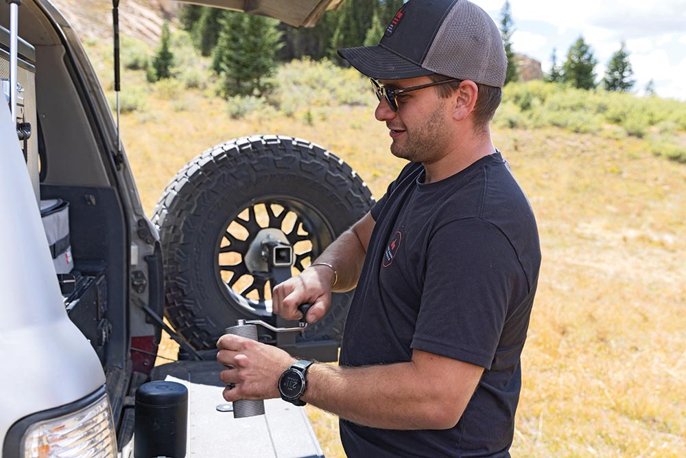 A man in sunglasses and a hat uses a hand-operated coffee bean grinder on the tailgate of the Land Cruiser.