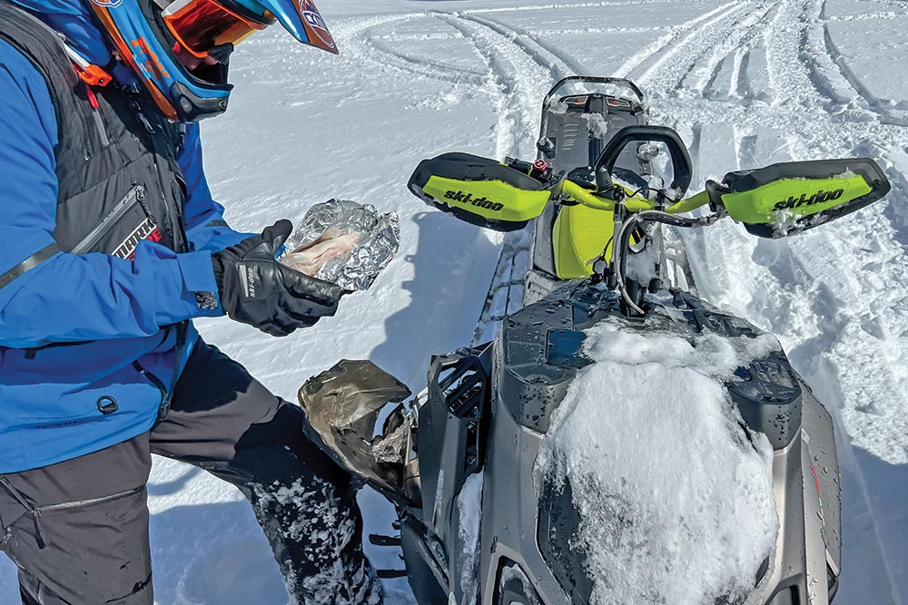 Rider holds a sandwich beside a ski-doo snow mobile. 