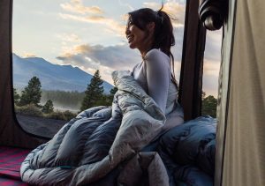 A woman wrapped in a blanket sits in a roof top tent.