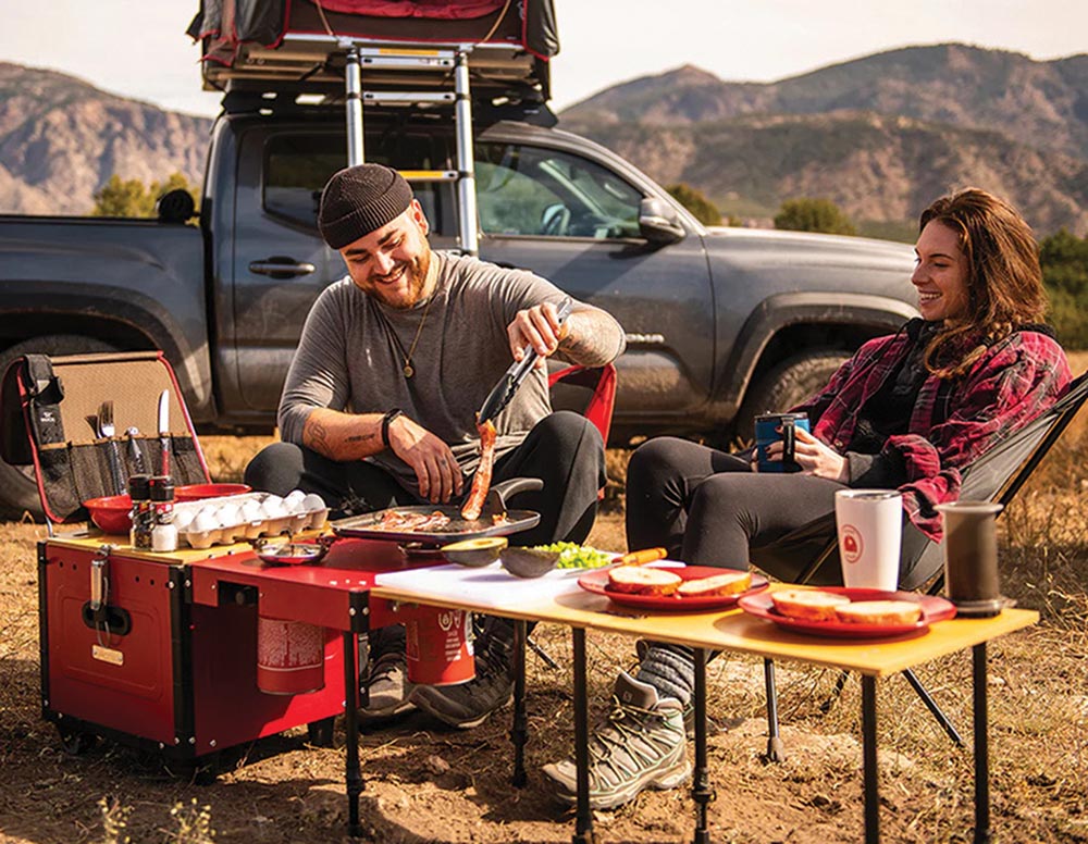 A man and a woman sit at the tables that fold out of the Aioks, the man is cooking food and the woman is drinking coffee.