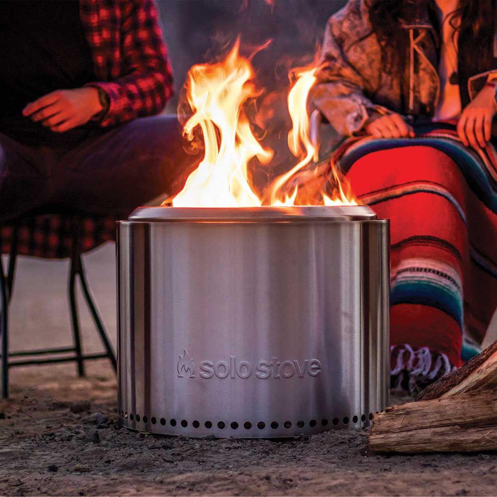 People in chairs with blankets on their laps gather around the cylindrival metal fire pit.