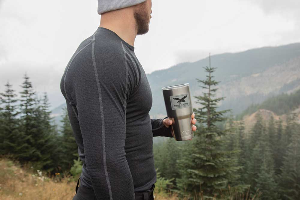 A man wearing a gray base layer shirt and holding a stainless tumbler stands in the woods.