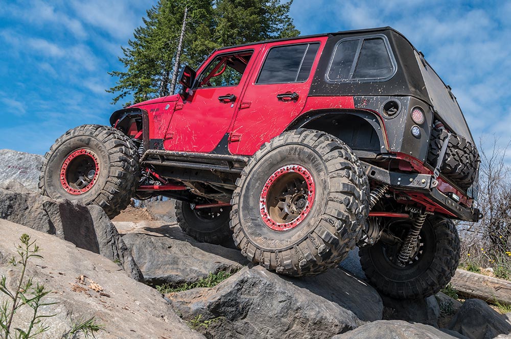 A modern red Jeep with large wheels and a black soft top climbs up rocks.