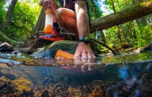 A man dips the water bottle into a stream while holding the filter.