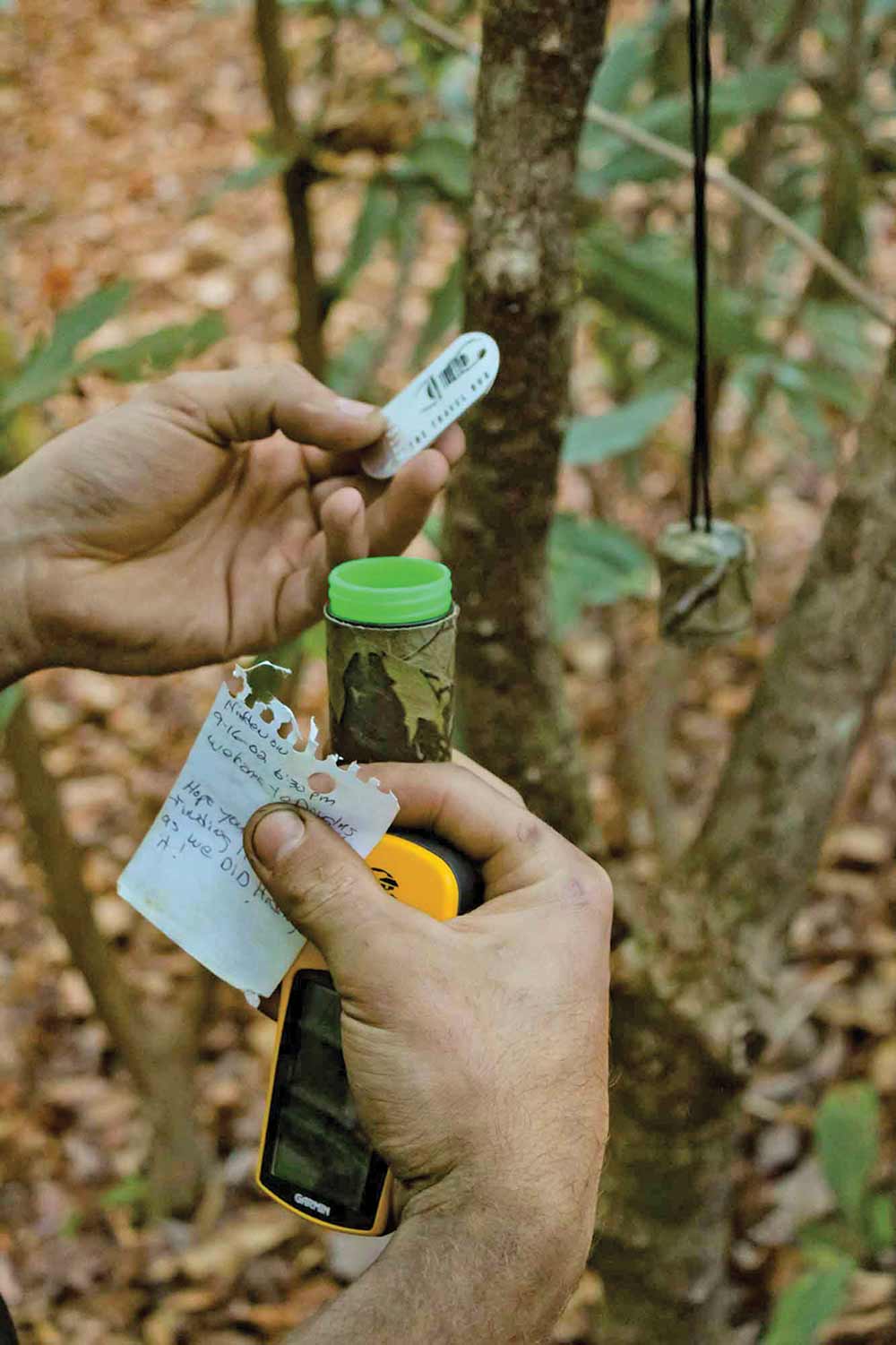 A geocacher opens a camo-cover cache hanging from a tree limb. Inside is a trackable dog tag and a note from the owner.